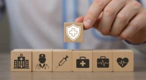 Hand placing a wooden block with a white medical shield and cross atop a row of healthcare-themed blocks.