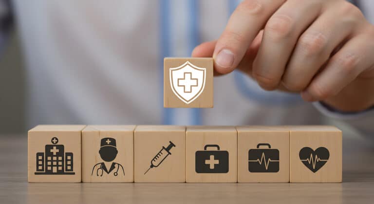 Hand placing a wooden block with a white medical shield and cross atop a row of healthcare-themed blocks.