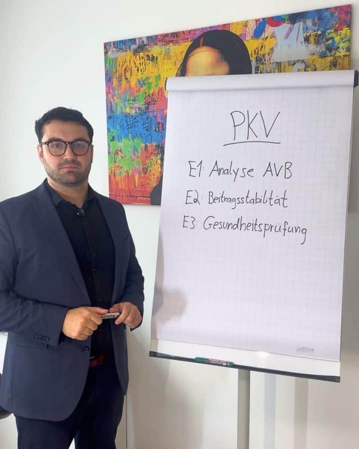 Man in a navy suit standing beside a flip chart with handwritten German notes: PKV, E1 Analyse AVB, E2 Beitragsstabilität, E3 Gesundheitsprüfung.